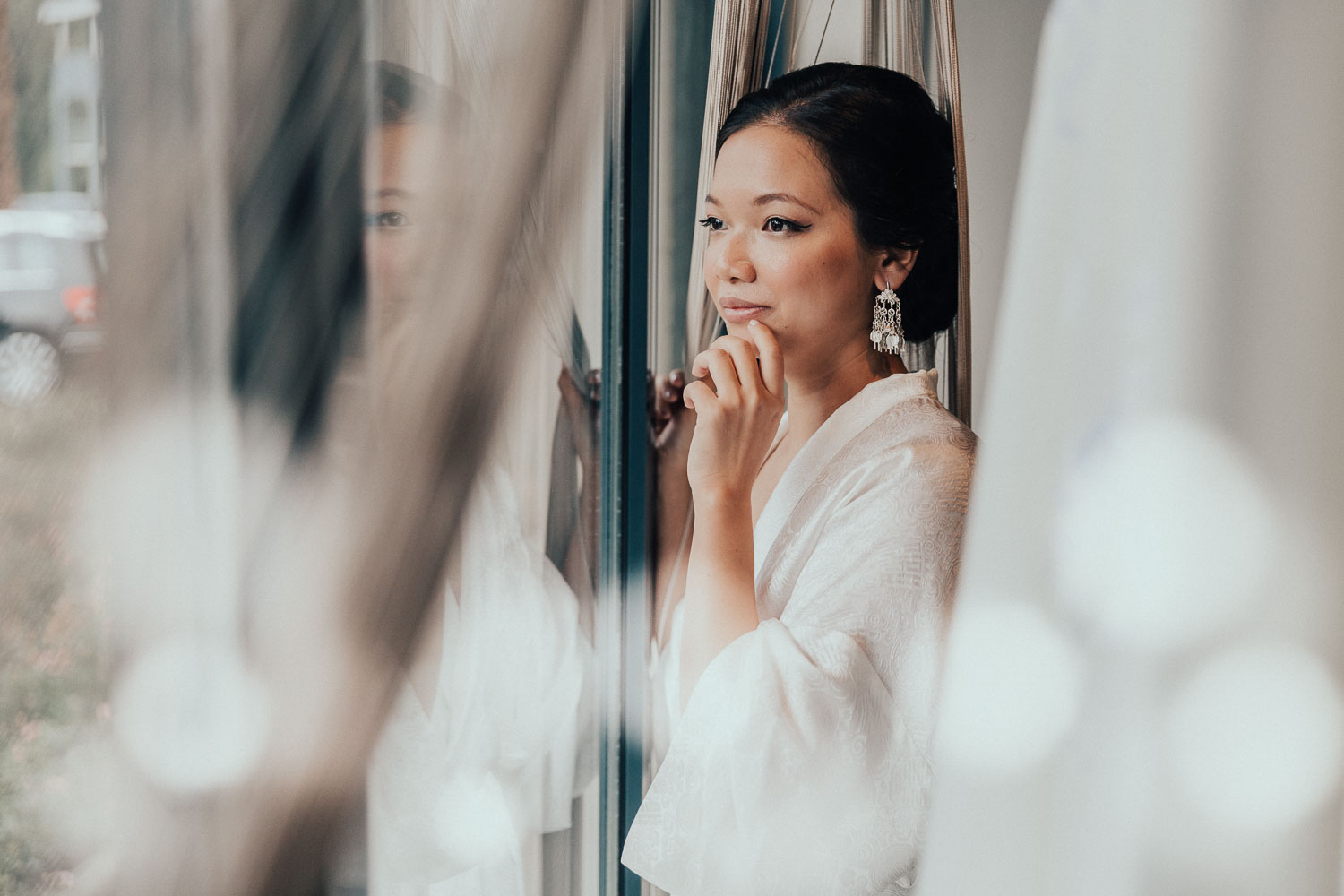 Aziatische bruid blijft dromerig bij de ramen en licht glimlachend terwijl de fotograaf in Amsterdam een spontane en openhartige foto maakt