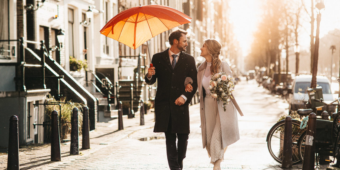 Echtpaar man en vrouw lopen met een rode paraplu terwijl trouwfotograaf in Amsterdam foto's maakt