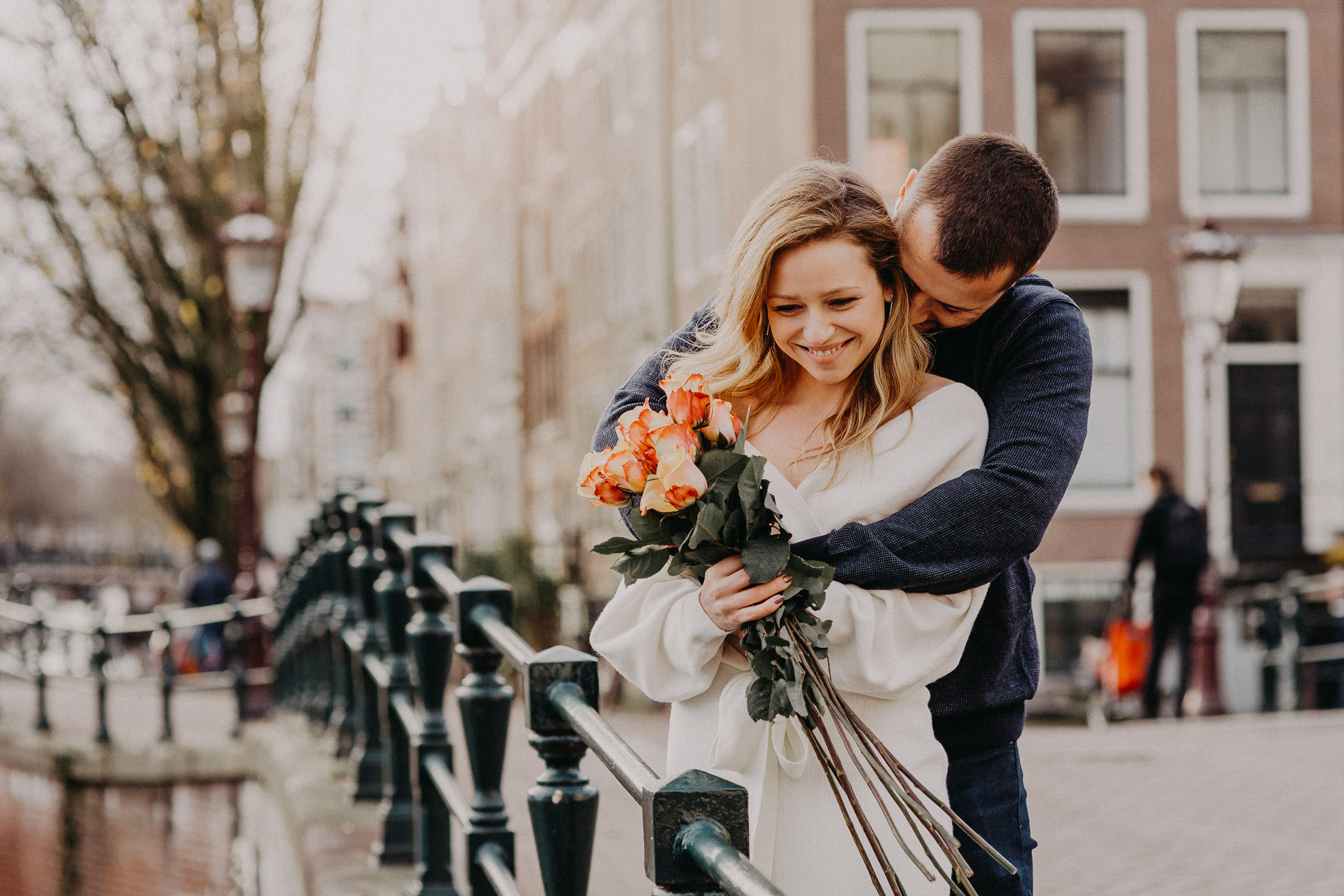 Married couple is posing on the bridge for a wedding photographer in Amsterdam. Man is hugging a woman from behind. She is smiling and carries orange roses.