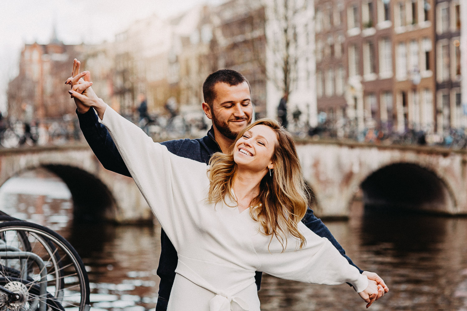 Een gelukkig mooi stel houdt de handen wijd vast tijdens een fotoshoot voor een trouwfotograaf in Amsterdam. Ze blijven voor de kanaalbrug in Amsterdam en glimlachen.