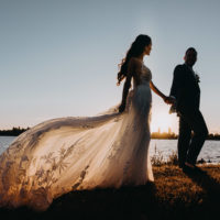 Een bruid in een trouwjurk met lange staart met een hand van een bruidegom. Ze lopen tijdens de zonsondergang op de kustlijn in Nederland. Stel poseert voor fotograaf in Amsterdam.