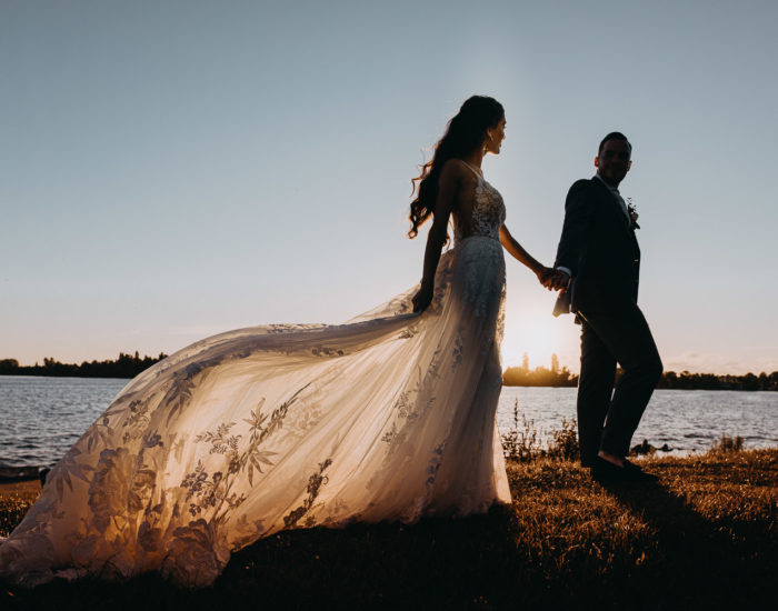 Een bruid in een trouwjurk met lange staart met een hand van een bruidegom. Ze lopen tijdens de zonsondergang op de kustlijn in Nederland. Stel poseert voor fotograaf in Amsterdam.