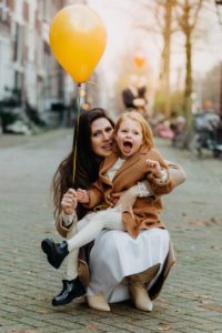 mom and daughter are posing for a family photo session in Amsterdam, mom is holding an orange balloon in her hands, and daughter is laughing