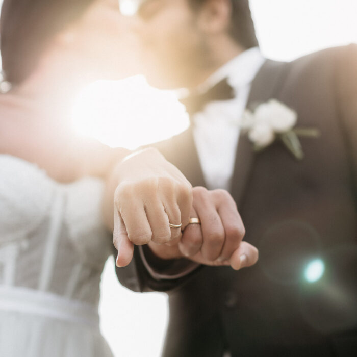 Close-up of a bride and groom showing their wedding rings during a golden hour portrait, with soft sunlight and lens flare creating a romantic atmosphere.