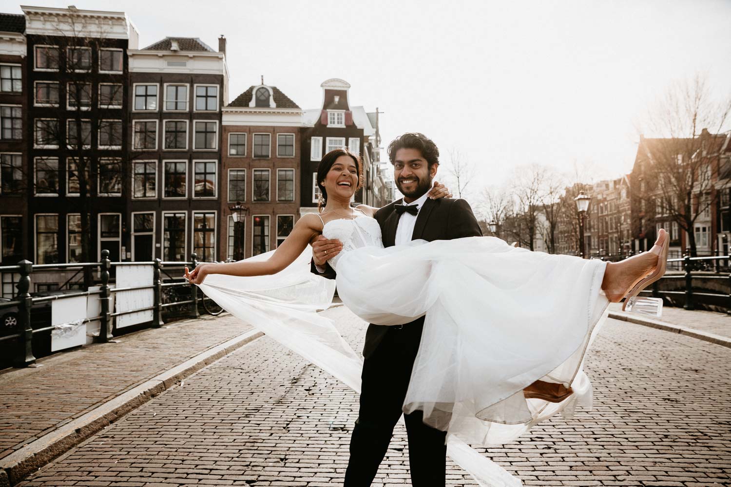 the couple stands on the pavement at the Indian wedding in Amsterdam. The groom in a black suit holds the bride in his arms. The bride is holding a bouquet of white roses.
