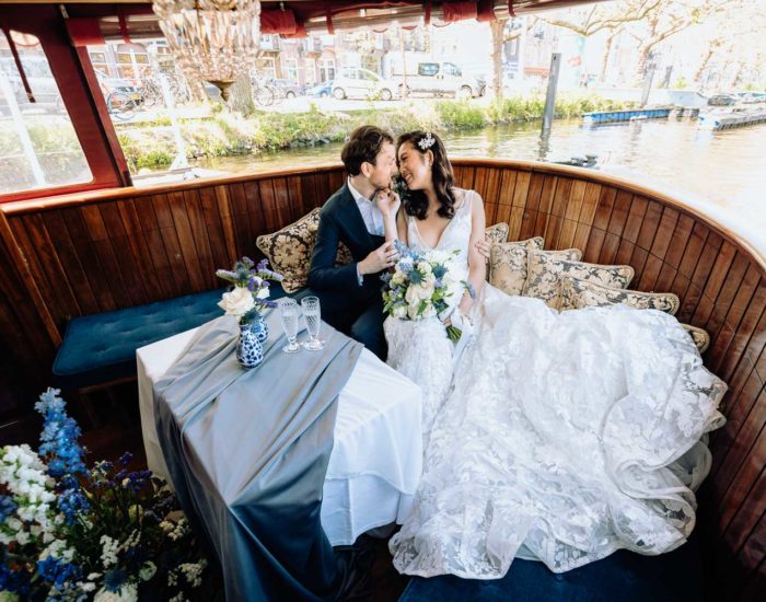 The newlyweds sit on the deck of the Soeverein boat on one of the Amsterdam canals, the bride holds a beautiful bouquet of white roses in her hands, and the groom looks at the bride