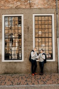 Gay couple at the gay wedding photoshoot staying in front of the old building with big windows and smiling each other.