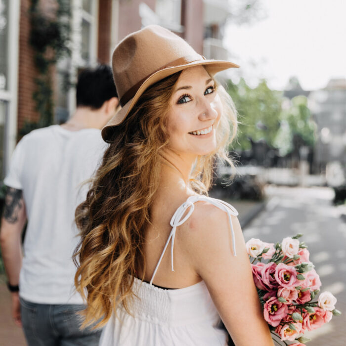 Woman holding a bouquet of pink flowers and smiling back while walking through a quiet Amsterdam street during a couples photoshoot.