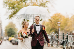 Bride and groom walking down the street under the umbrella during their autumn wedding photoshoot in Amsterdam. Photographer captures this moment of Amsterdam elopement. How to getting married in Amsterdam click on the photo.