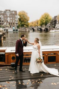 Groom kissing the hand of the bride in front of canal, while photographer makes photos. How to getting married in Amsterdam click on the photo and read our article