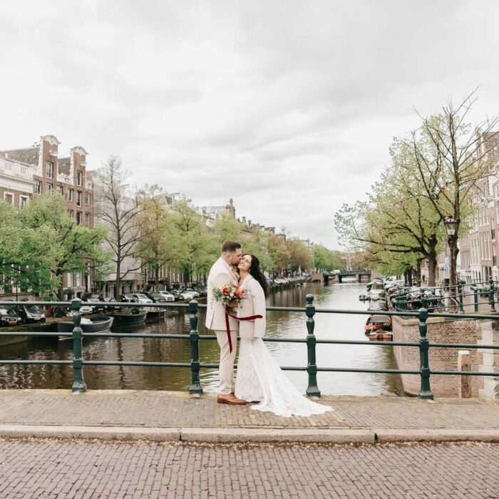 Bride and groom embracing on an Amsterdam canal bridge with historic canal houses and waterways creating a classic city backdrop.