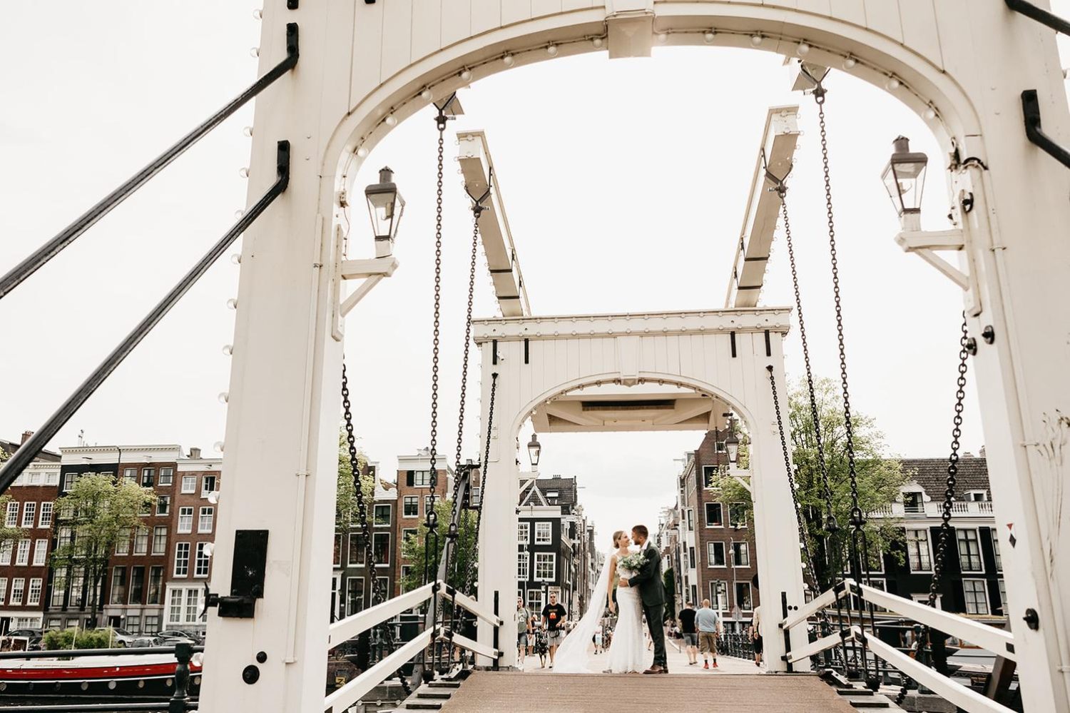Bride and groom sharing a kiss beneath Amsterdam's iconic Magere Brug, with historic canal houses and the city's famous drawbridge framing the scene.
