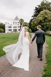 Romantic wedding at Oranjerie Landgoed Sandenburg, featuring a newlywed couple walking hand in hand towards a stunning white castle. The bride’s elegant long veil flows gracefully, adding a dreamy touch to this luxury wedding location in the Netherlands. A perfect setting for an exclusive wedding venue with a fairytale charm.
