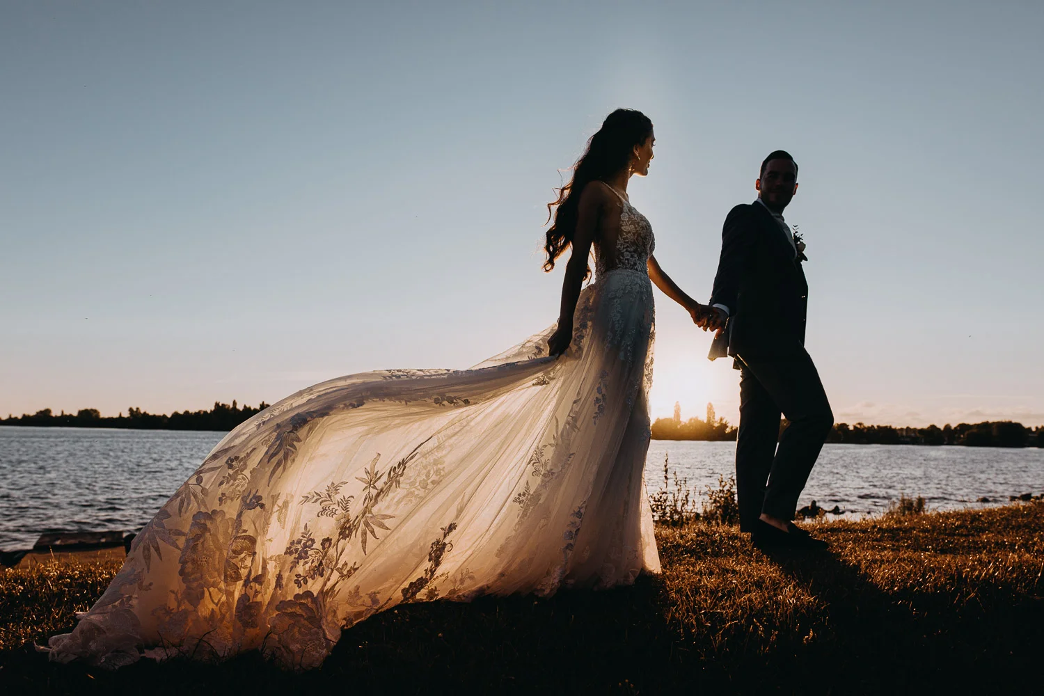 A bride in a long tail wedding dress holding a hand of a groom. They walking on the costline in Netherlands during the sunset time. Couple is posing for photographer in Amsterdam.